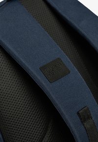 Sangle de sac à dos en tissu bleu avec rembourrage en maille noire et une étiquette noire cousue estampillée "SUPER DRY®".