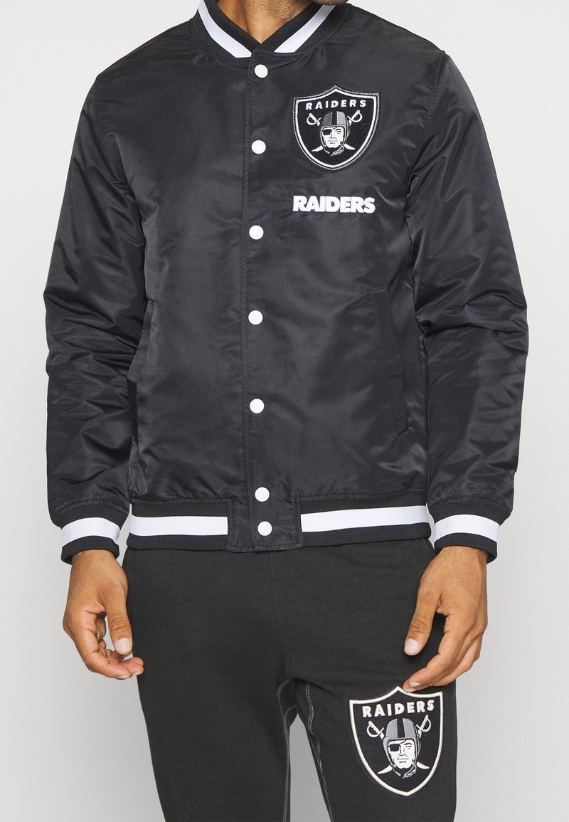 Černá bomber bunda s bílými zapínacími knoflíky, obsahuje logo Raiders na hrudi a pruhované žebrované manžety a pás.