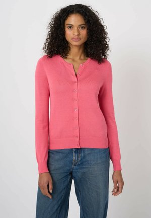 Kobieta o kręconych włosach, ubrana w zapinany na guziki różowy kardigan i niebieskie dżinsy, stojąca na tle jednolitego, jasnego tła.