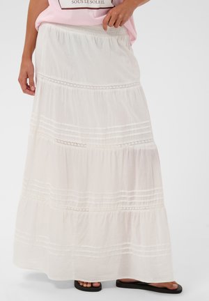 Jupe longue blanche à volants avec bordure en dentelle portée avec une chemise rose et des sandales noires à bout ouvert, main reposant sur la taille.