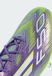 Paarse voetbalschoen met een gestructureerd oppervlak. Kenmerkt zich door contrasterende groene accenten en witte branding, samen met paarse veters en een gestroomlijnd ontwerp.