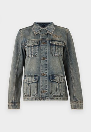 Person trägt Sonnenbrille, Jeansjacke, weißes Oberteil, hellbeige Hose, schwarze Schuhe, trägt große braune Wildledertasche auf der Schulter vor weißem Hintergrund.