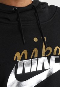 Svart hoodie med svarta dragsko, med metallisk guld kursiv text delvis täckt av en stor silver Nike-logotyp och swoosh.