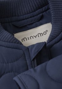 Navy modra jakna z uleknjeno zasnovo, rebrastim ovratnikom in zadrgo. Oznakovana z "minymo" na svetli tkaninski oznaki.