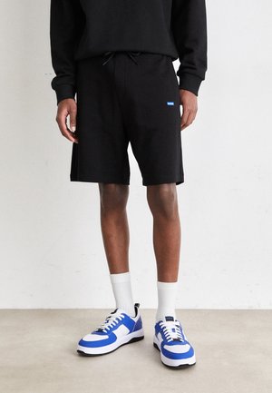 Person iført sorte shorts med et lille blåt logo, hvide crew-sokker og hvide og blå atletiksko, står på et lyst gulv.