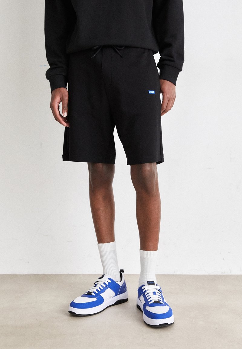 Personne portant un short noir avec un petit logo bleu, des chaussettes blanches et des baskets blanches et bleues, debout sur un sol clair.