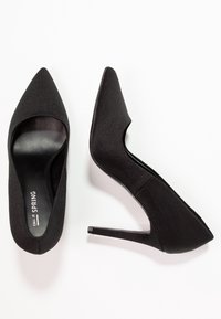 Chaussures à talons hauts noires avec un bout pointu, une tige en tissu lisse et un talon stiletto. Intérieur doté d'une doublure noire lisse, label de la marque visible.