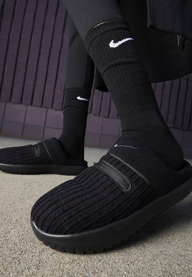 Zapatos negros tipo slip-on con parte superior de tela acanalada y suela de goma, combinados con calcetines negros hasta la rodilla que presentan un logo blanco. Superficie del suelo visible.