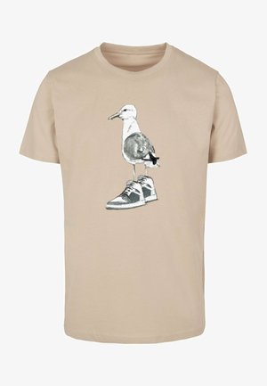 Bežinė medvilninė marškinė su atvaizdu, kuriame pavaizduota žuvėdra, dėvinti sportinius batelius, su monochrominėmis detalėmis ir laisvu siluetu.