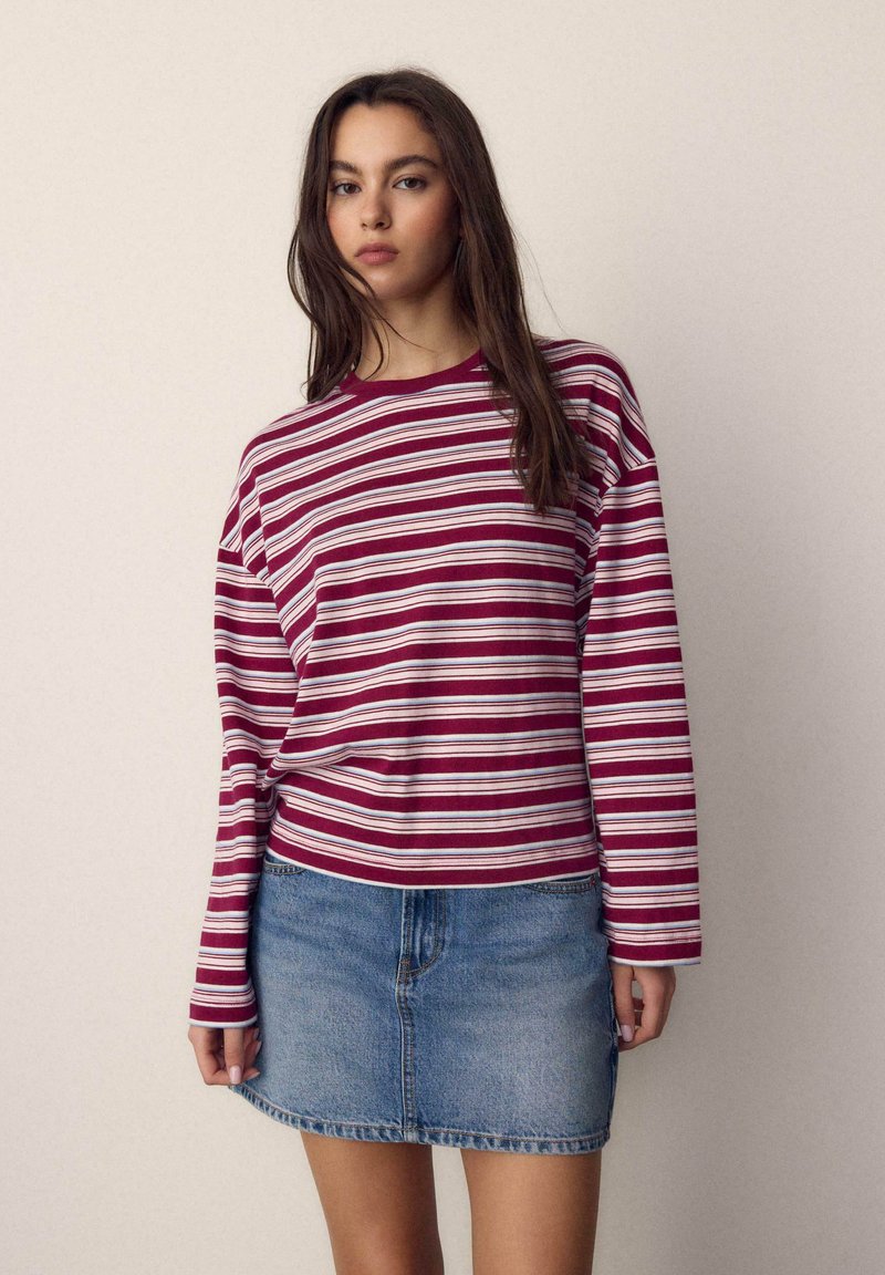 Jeune femme aux longs cheveux bruns portant un t-shirt à manches longues rayé bordeaux et blanc ainsi qu'une mini-jupe en jean bleue, se tenant devant un fond uni.
