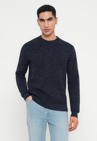 Man wearing a dark blue crewneck sweater and light blue jeans, standing with one hand in pocket against a plain white background.