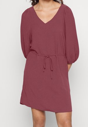 Vrouw draagt een knielange, lange mouwen mauve jurk met een V-halslijn en een taille met trekkoord.