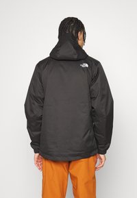 Svart vattentät jacka med huva, som har elastiska ärmslut och en logotyp på höger axel. Bärs med orange byxor.