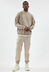 Brun sweatshirt med ribbade mudd och nederkant, kombinerad med ljusbeige cargobyxor med sidofickor och elastiska fotled. Vita högsneakers.