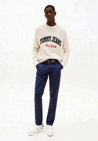 Young man wearing a white "Tommy Jeans New York" sweater, navy blue pants with a black belt, and white sneakers, standing with hands in pockets.