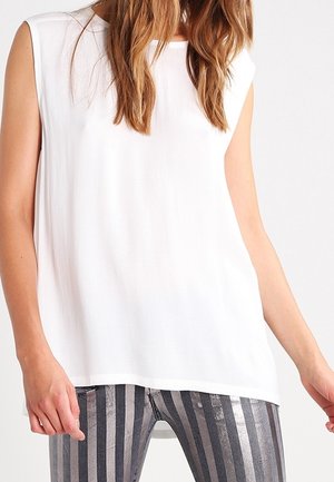 Mouwloze witte blouse gedragen met metallic verticaal gestreepte leggings, waarbij de torso en armen zichtbaar zijn tegen een effen achtergrond.