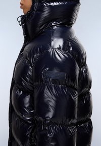 Giacca puffer nera lucida con colletto alto e trapuntatura pronunciata. Presenta un patch con il marchio sulla manica e una tasca con zip.