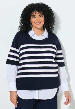Vrouw met krullend zwart haar draagt een marineblauw-wit gestreepte korte mouwen trui over een wit geknoopt overhemd, glimlachend tegen een witte achtergrond.