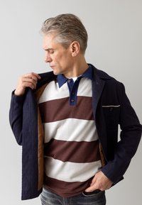 Navy textured jacket over a brown and white striped polo shirt. Jacket features a brown interior and small chest pocket. Casual denim jeans.