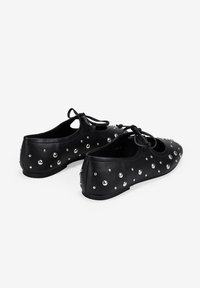 Zapatillas de ballet planas de cuero negro con tachuelas redondas plateadas y cordones frontales anudados, mostradas desde ángulos trasero y lateral sobre un fondo blanco.