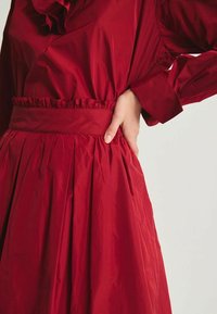 Robe rouge avec un haut froncé, une taille cintrée et une jupe plissée. Fabriquée en tissu léger avec une texture lisse.