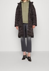 Manteau long noir matelassé avec capuche, porté sur un sweat à capuche vert clair, associé à un jean gris évasé et des mules noires à talons blocs.