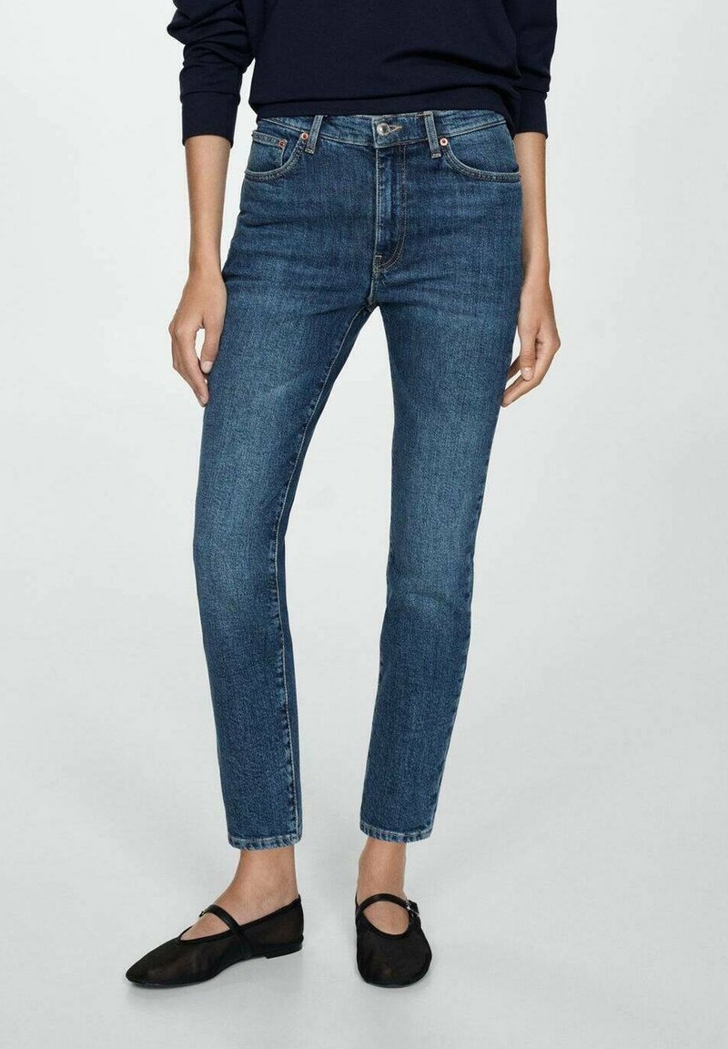Femme portant un jean bleu slim, un haut noir à manches longues et des ballerines Mary Jane noires, debout devant un fond clair uni.