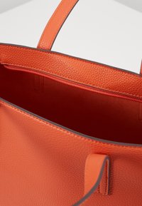 Pomarańczowa teksturowana torba tote z wewnętrzną kieszenią na zamek, gładkimi uchwytami i kontrastującym przeszyciem wzdłuż krawędzi.