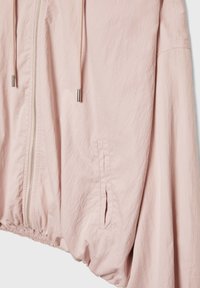 Jasnoróżowa lekka kurtka z przednim zamkiem błyskawicznym, kapturem ze sznurkiem, elastycznym dołem i bocznymi kieszeniami. Materiał o fakturze z miękkim wykończeniem.