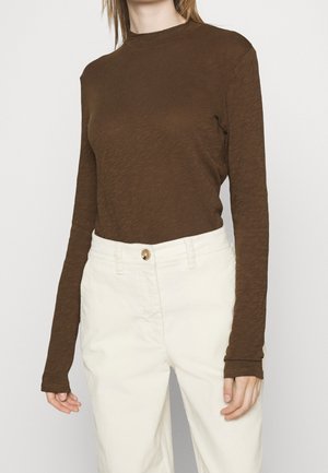 Femme portant une chemise marron à manches longues rentrée dans un pantalon taille haute écru avec une fermeture à boutons.
