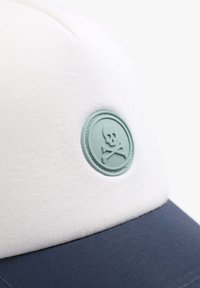 Gorra con una parte superior de tela blanca y un visera azul marino, con una insignia redonda de goma verde menta que muestra un diseño de calavera y tibias cruzadas.