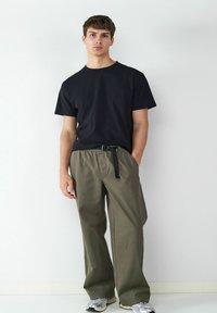 Zwarte T-shirt met ronde hals gecombineerd met losse olijfgroene cargobroek met een zwarte riem. Model draagt grijze sneakers.