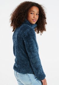 Maglione a collo alto in pile blu, morbido e fluffoso, con una superficie texturizzata. Il capo ha un design aderente e maniche lunghe, abbinato a jeans in denim chiaro.