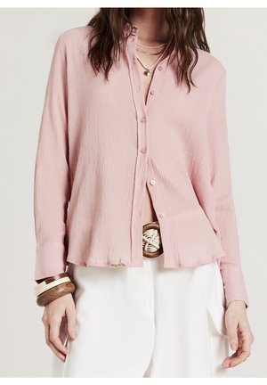 Femme portant une chemise boutonnée texturée rose pâle, un pantalon blanc avec une grande boucle de ceinture ronde, des bracelets en bois et des colliers superposés.