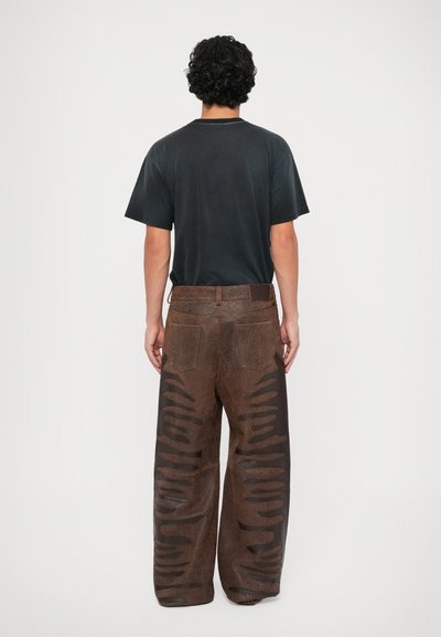 Bruine leren broek met uitsparingen, wijd lopend ontwerp en twee achterzakken. Gecombineerd met een zwart T-shirt, gezien vanuit de achterkant.