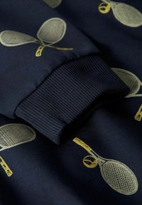 Tamsiai mėlynas džemperis su tekstūruotu rankogaliu. Audinys puoštas šviesiai geltonų teniso raketų ir kamuoliukų spaudiniu, kuris sukuria žaismingą raštą.