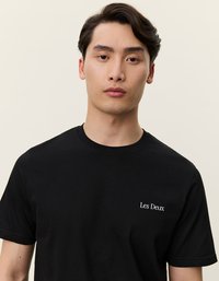 Μαύρη βαμβακερή μπλούζα με στρογγυλή λαιμόκοψη και κοντά μανίκια, με λευκό κεντημένο κείμενο "Les Deux" στην αριστερή πλευρά του στήθους.