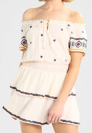 Beige schulterfreie Bluse mit blau-roten, bestickten Blumenmustern, ausgestattet mit einer Vorderbindetie und einem gerafften Taillenbund, kombiniert mit einem mehrlagigen Rock.