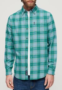 Camicia a quadri verde e blu con colletto, dotata di taschino sul petto e di un etichetta sull'orlo. Realizzata in un tessuto morbido.