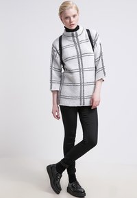 Hvit genser med svart rutemønster, høy krage og trekvart ermer, kombinert med svarte leggings og svarte snørestøvler.