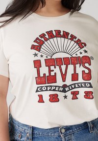 Osoba v bílém tričku Levi's s červeným a černým textem "Authentic Levi's Copper Riveted 1873", doplněná modrými džíny.
