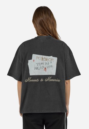 Person mit langen braunen Haaren, trägt ein schwarzes übergroßes T-Shirt mit einem verblassten Ticketmotiv und dem Schriftzug "Moments to Memories" auf der Rückseite.