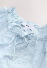 Camicia azzurro chiaro in tessuto strutturato, con dettagli arricciati, un fiocco al collo e un'etichetta con la scritta "NEXT".