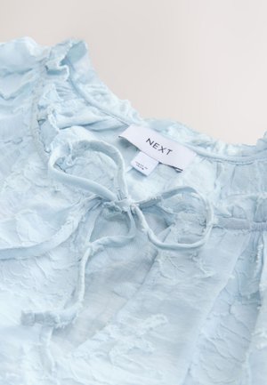 Bluza bleu deschis din material texturat, cu accente volanate, detaliu de legare la gât și etichetă inscripționată cu "NEXT".