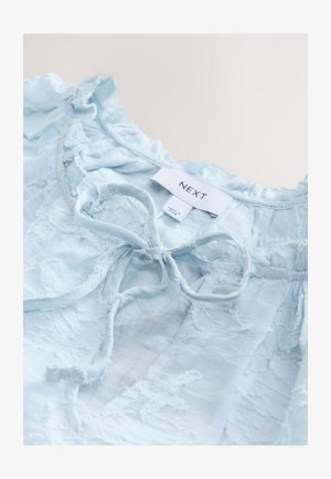 Bluza bleu deschis din material texturat, cu accente volanate, detaliu de legare la gât și etichetă inscripționată cu "NEXT".