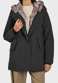 Svart parkas med huva i fuskpäls, dragkedja framtill, två sidofickor och avslappnad passform. Slät yttertyg, mjukt inre material.