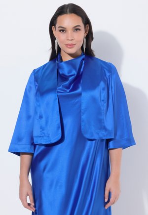 Mujer con cabello largo y oscuro que lleva un vestido azul satinado brillante y una chaqueta corta a juego, complementado con largos pendientes plateados, sobre un fondo blanco.