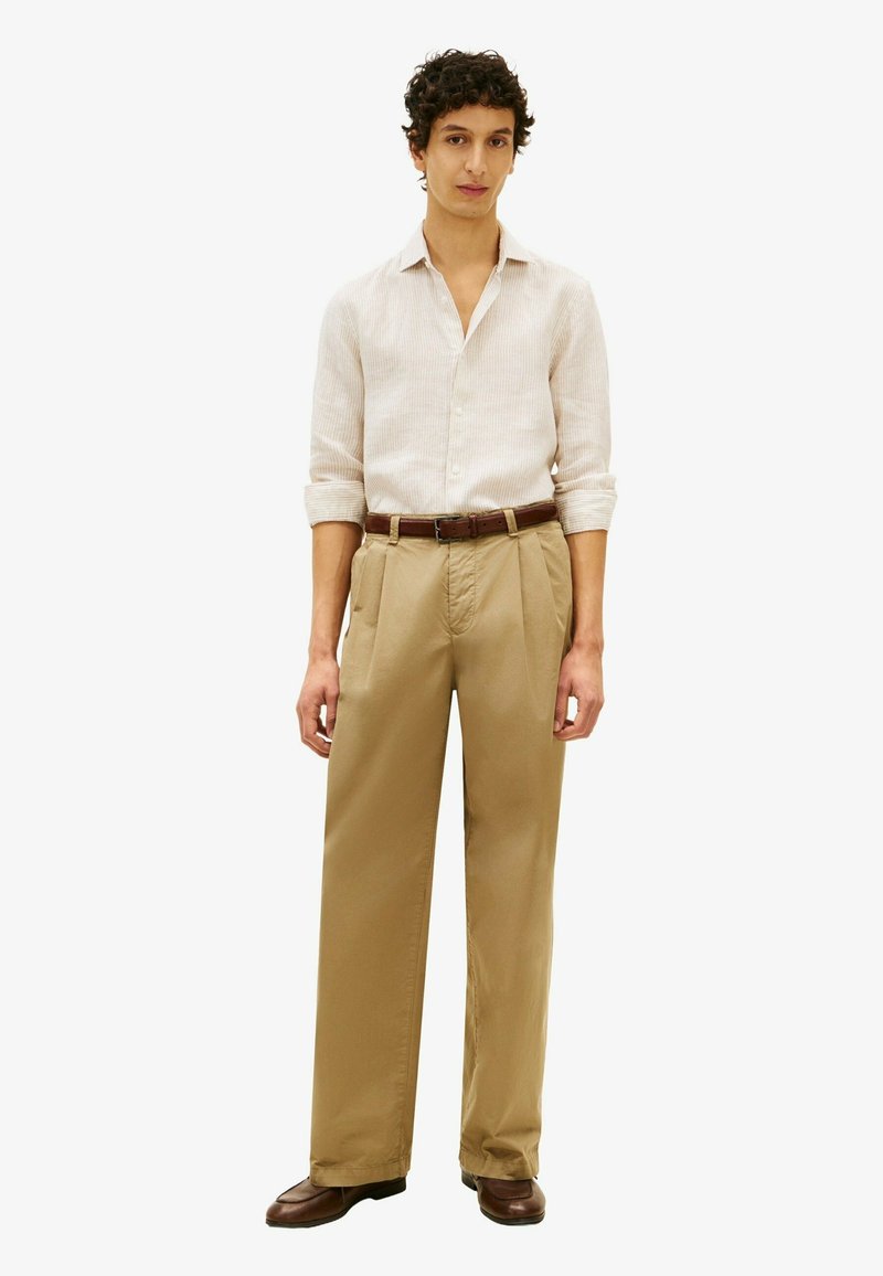 Homme portant une chemise à manches longues rayée beige clair, rentrée dans un pantalon kaki taille haute à jambes larges avec une ceinture marron et des chaussures marron.