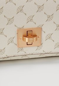 Cream leather surface with a textured pattern featuring geometric shapes; bronze lock closure with engraved branding detail.