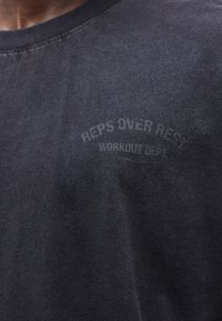 Gros plan d'un t-shirt gris foncé avec le texte délavé "REPS OVER REST WORKOUT DEPT." sur la zone gauche de la poitrine.